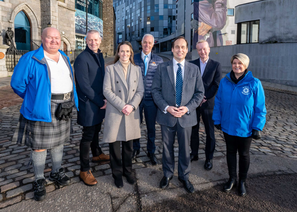 Chris Foy (CEO, VisitAberdeenshire),Rosemary Michie (Manager, Charles Michie Pharmacy), Bob Sanguinetti (CEO, Port of Aberdeen), Councillor Martin Greig (Chair of Aberdeen's Organising Committee for Tall Ships 2025) and Adrian Watson (CEO, Aberdeen Inspired), flanked by two blue-coated VisitAberdeenshire volunteers